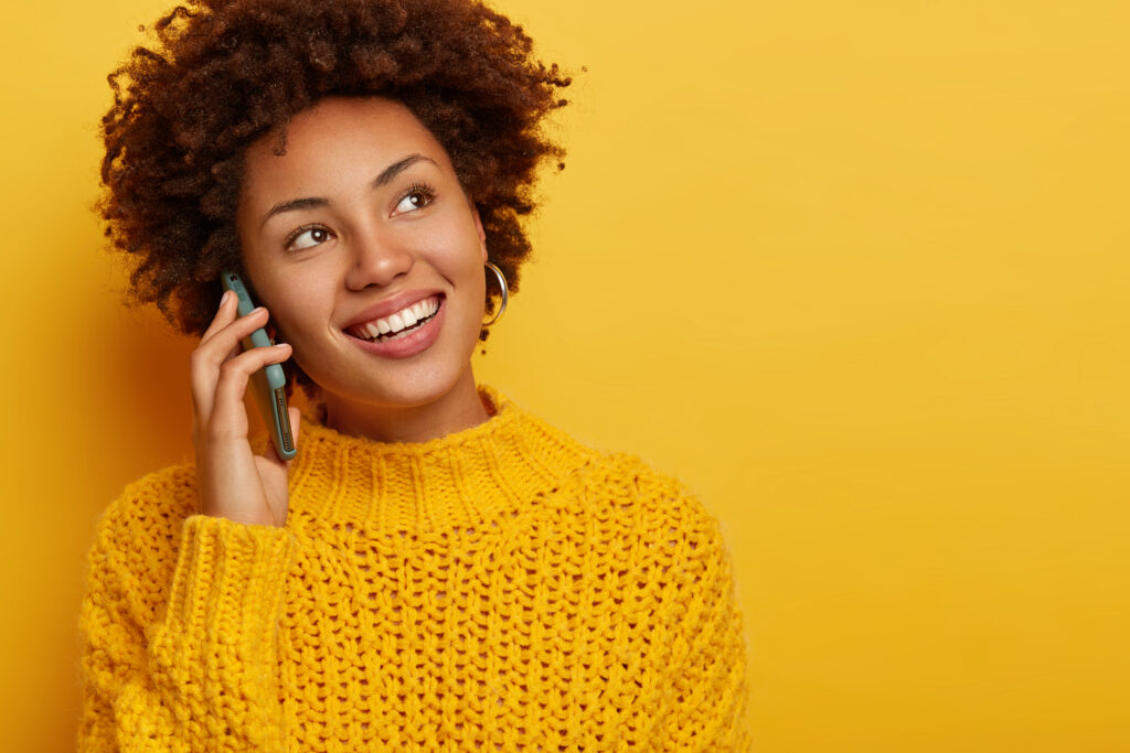 portrait of happy curly woman calls friend, enjoys pleasant conversation, holds mobile phone near ear, stays in touch, looks aside with broad toothy smile, wears knitted yellow jumper, blank space