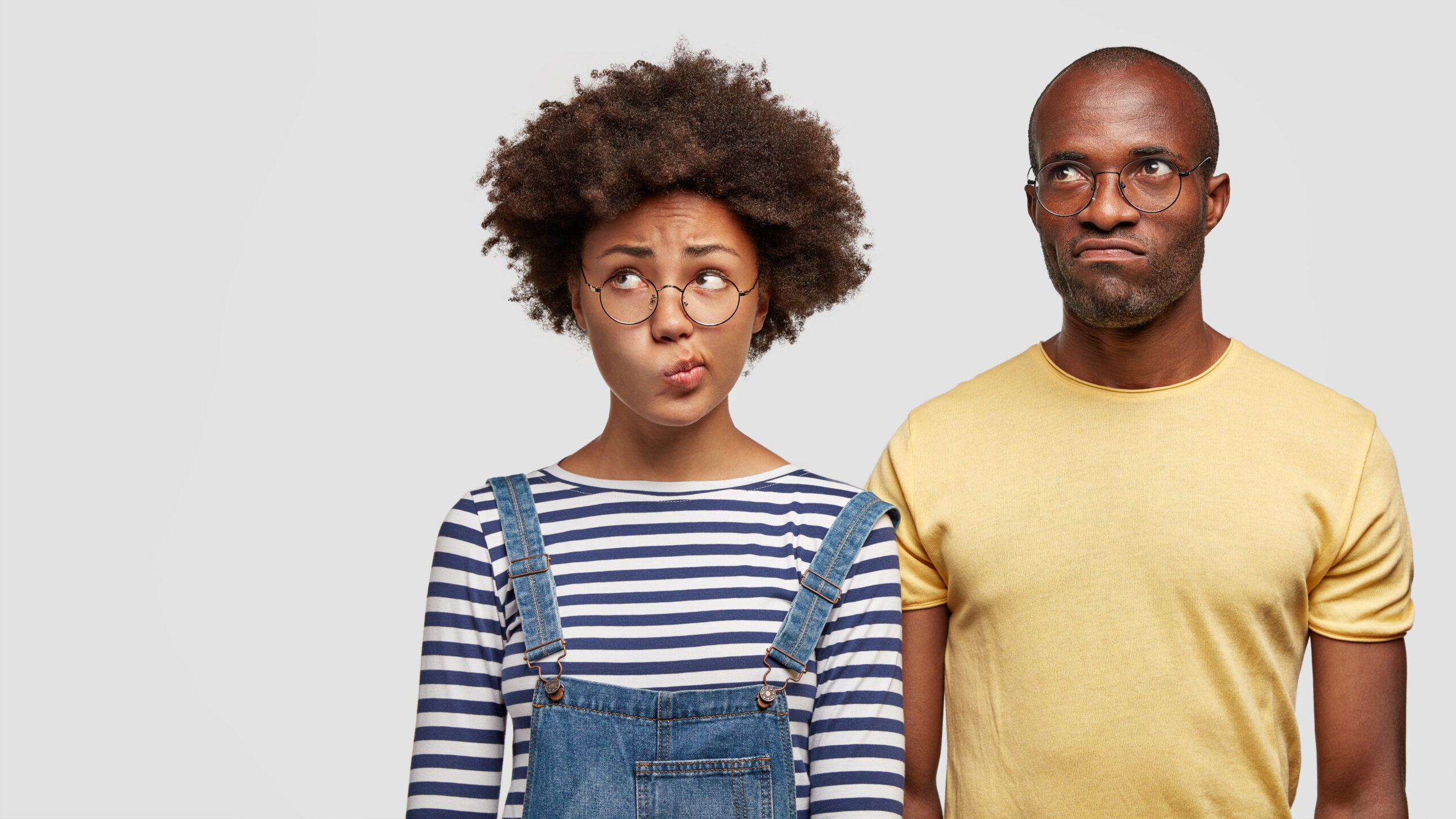 puzzled african american sister and brother curve lips and look with hesitation somewhere aside, can`t make decision. cute dark skinned woman in denim overalls spends free time with husband.