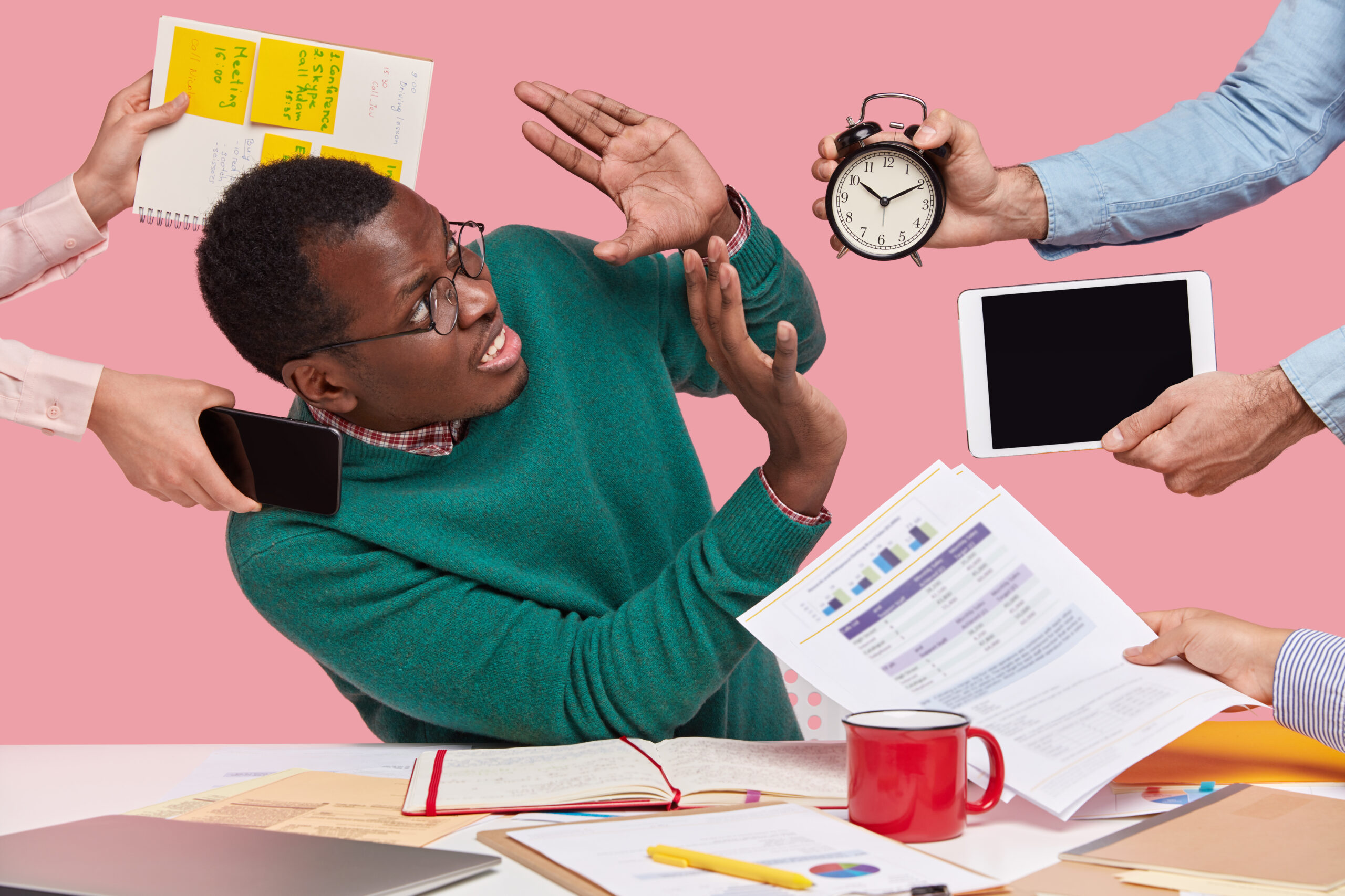 stressful dissatisfied dark skinned guy makes refusal sign, raises palms over alarm clock, touchpad and papers, has many tasks in one minute, poses at desktop, isolated over pink background. oh no!