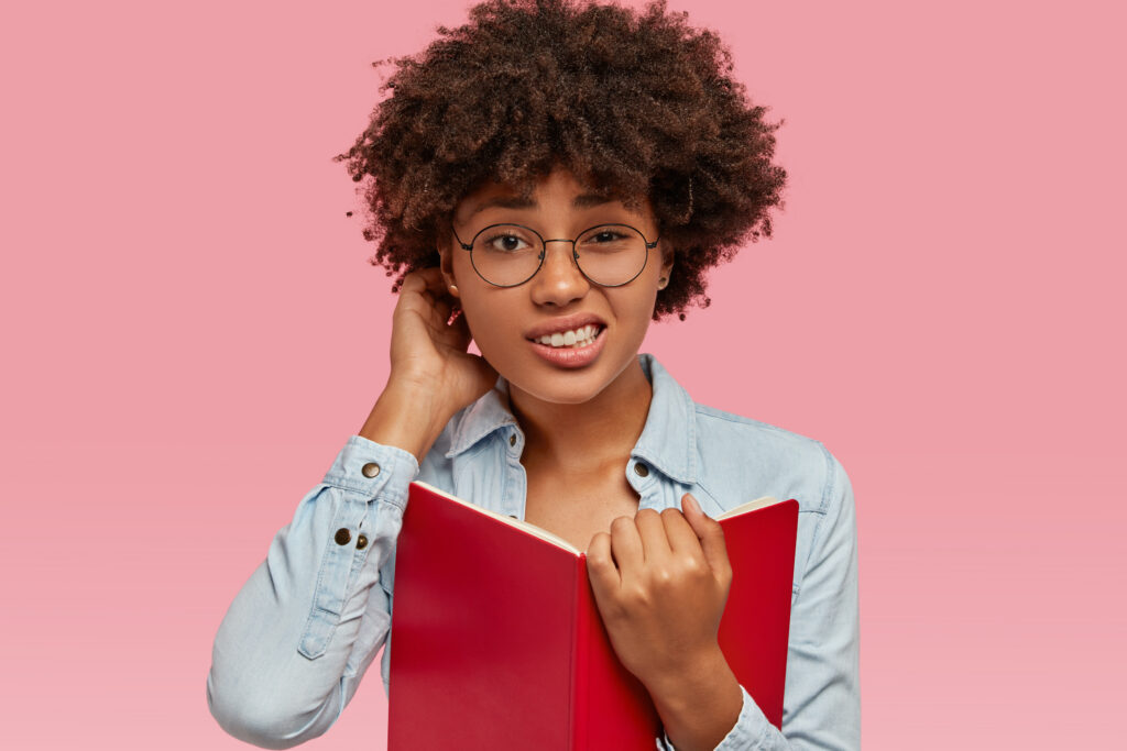 puzzled african american woman scratches head in bewilderment, cant remember important material and give answer on question immediately, holds red book in front, dressed in fashionable jacket